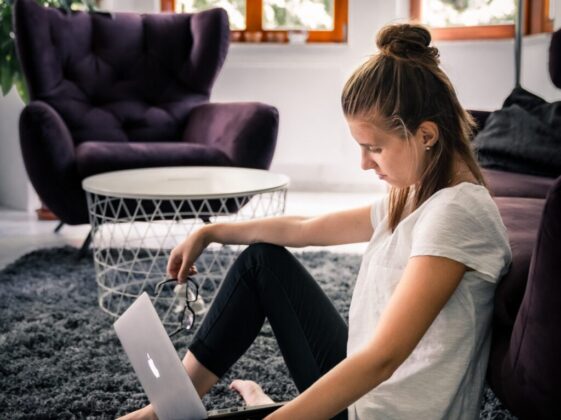 woman in white t-shirt and black leggings sitting on area rug while facing silver MacBook