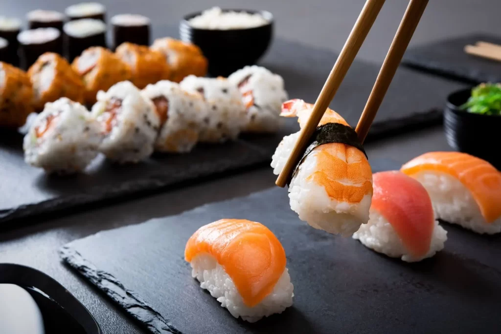 Krevetové sushi nigiri na černém talíři s dřevěnými hůlkami. Detailní záběr na nigiri s hůlkami a uramaki a hosomaki v pozadí